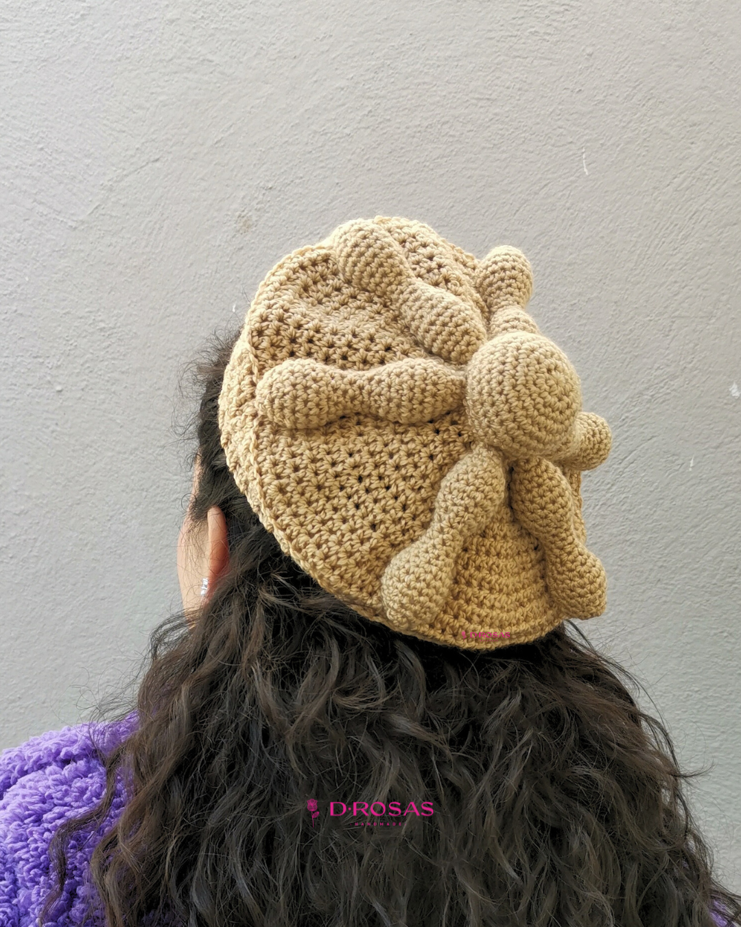 Boina en forma de pan de muerto tejida en crochet o gancho, color pan detalles en relieve que asemejan la forma del pan representativo de día de muertos. Usada por una mujer de cabello negro ondulado negro que usa un abrigo morado, vista de espaldas con la cabeza un poco hacia el lado.