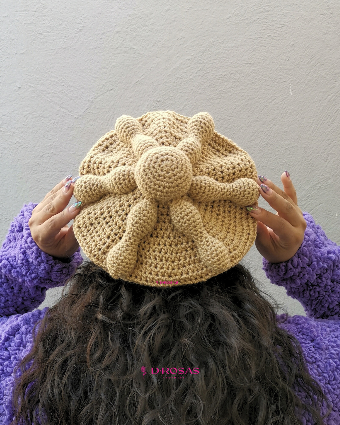 Boina en forma de pan de muerto tejida en crochet o gancho, color pan detalles en relieve que asemejan la forma del pan representativo de día de muertos. Usada por una mujer de cabello negro ondulado negro que usa un abrigo morado, vista de espaldas y agarrand con las manos la orilla de la boina.